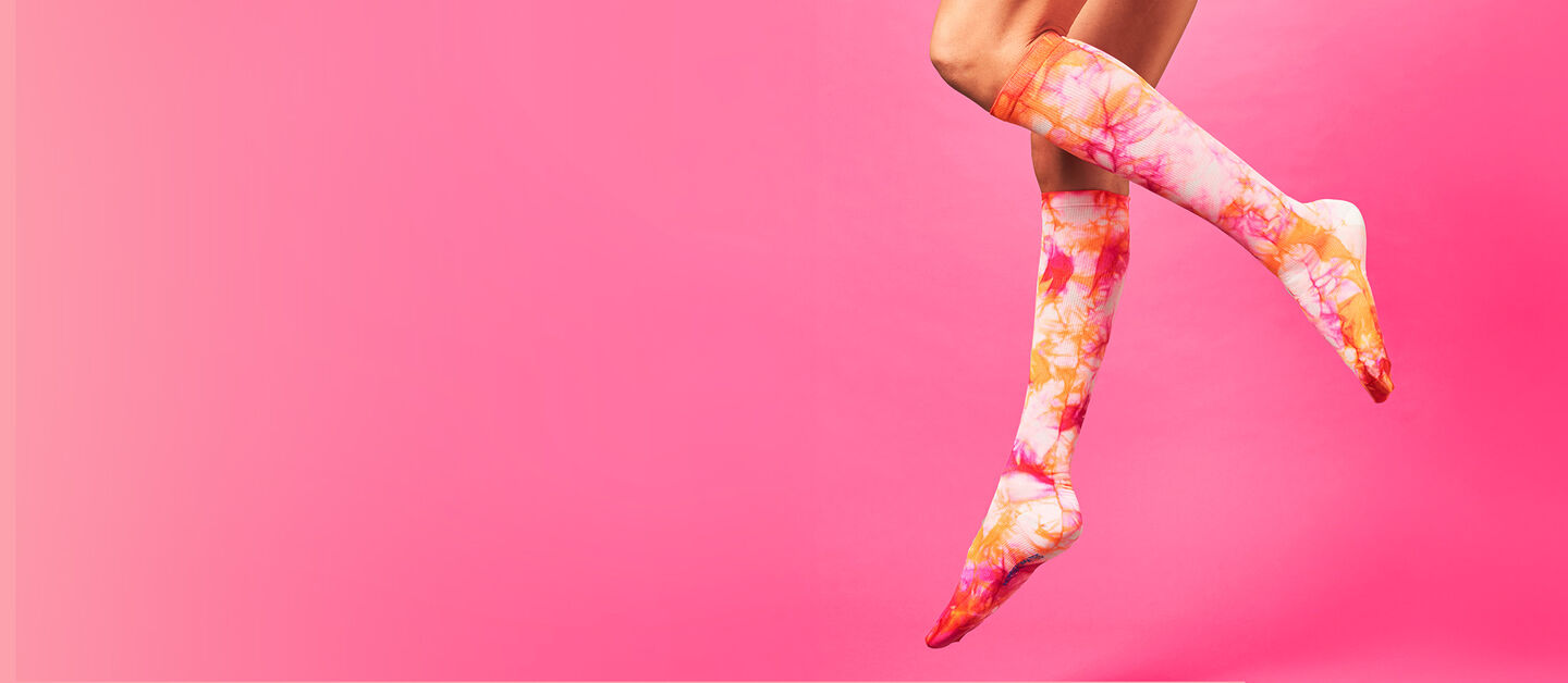 Fuchsia Flame pink and orange tie dye compression socks on woman’s legs, suspended in the air on a pink background